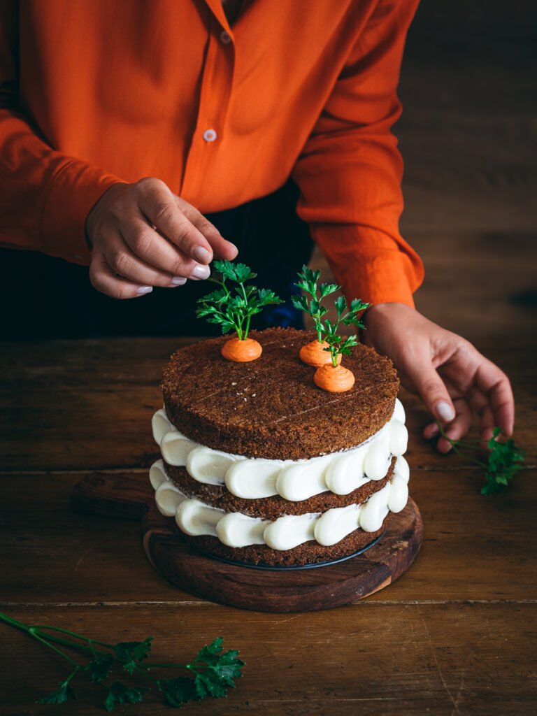 torta di carote e mele a strati con decorazione di frosting bianco e finte carote su tavolo legno