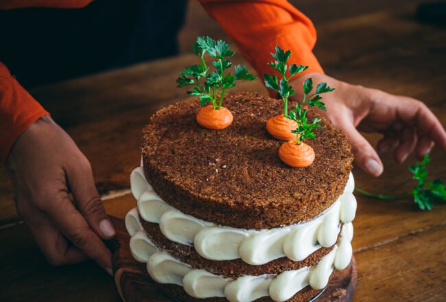 torta di carote e mele a strati con decorazione di frosting bianco e finte carote su tavolo legno