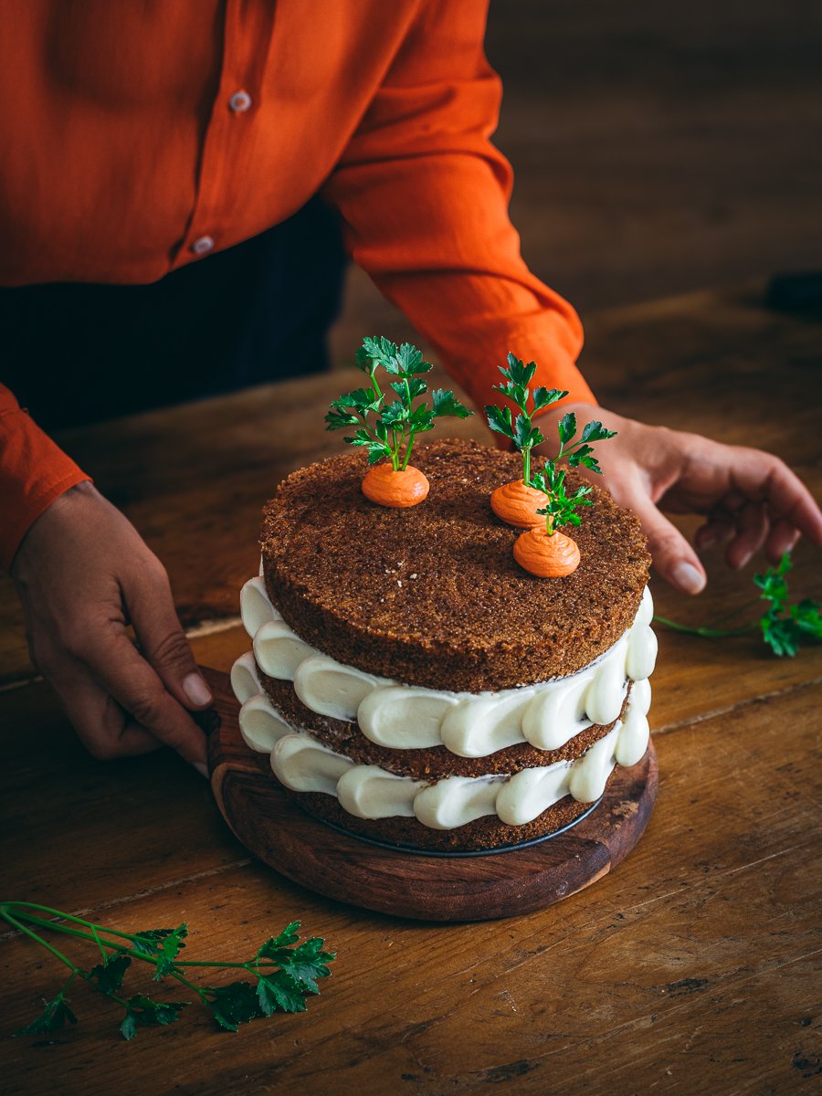 torta di carote e mele a strati con decorazione di frosting bianco e finte carote su tavolo legno